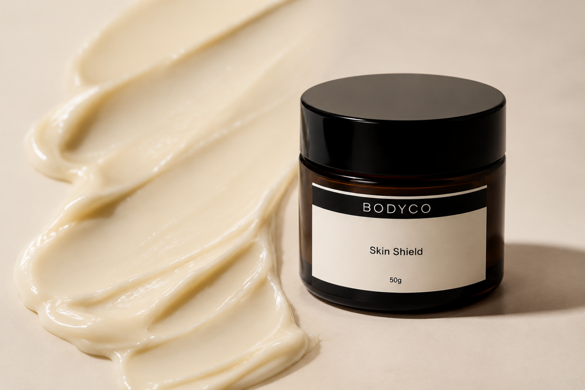 A dark amber glass jar of BODYCO Skin Shield sunscreen with a black lid and cream label, set next to a thick swatch of the beige cream product on a warm beige background.