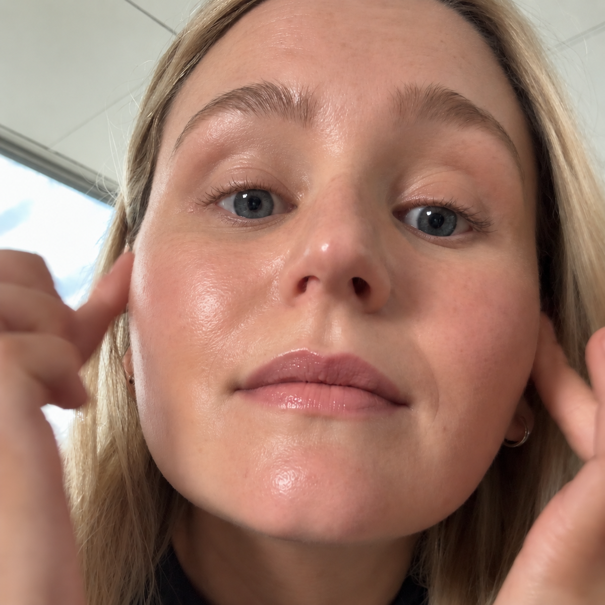 A close-up selfie of a woman with blonde hair and blue eyes, touching her ears with both hands, showing a clear view of her skin texture and makeup.
